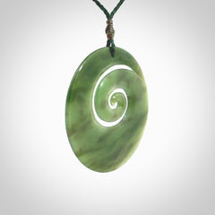 This picture shows a large hand-carved koru. It is carved from British Columbian jade and is suspended from a hand-plaited, green cord.