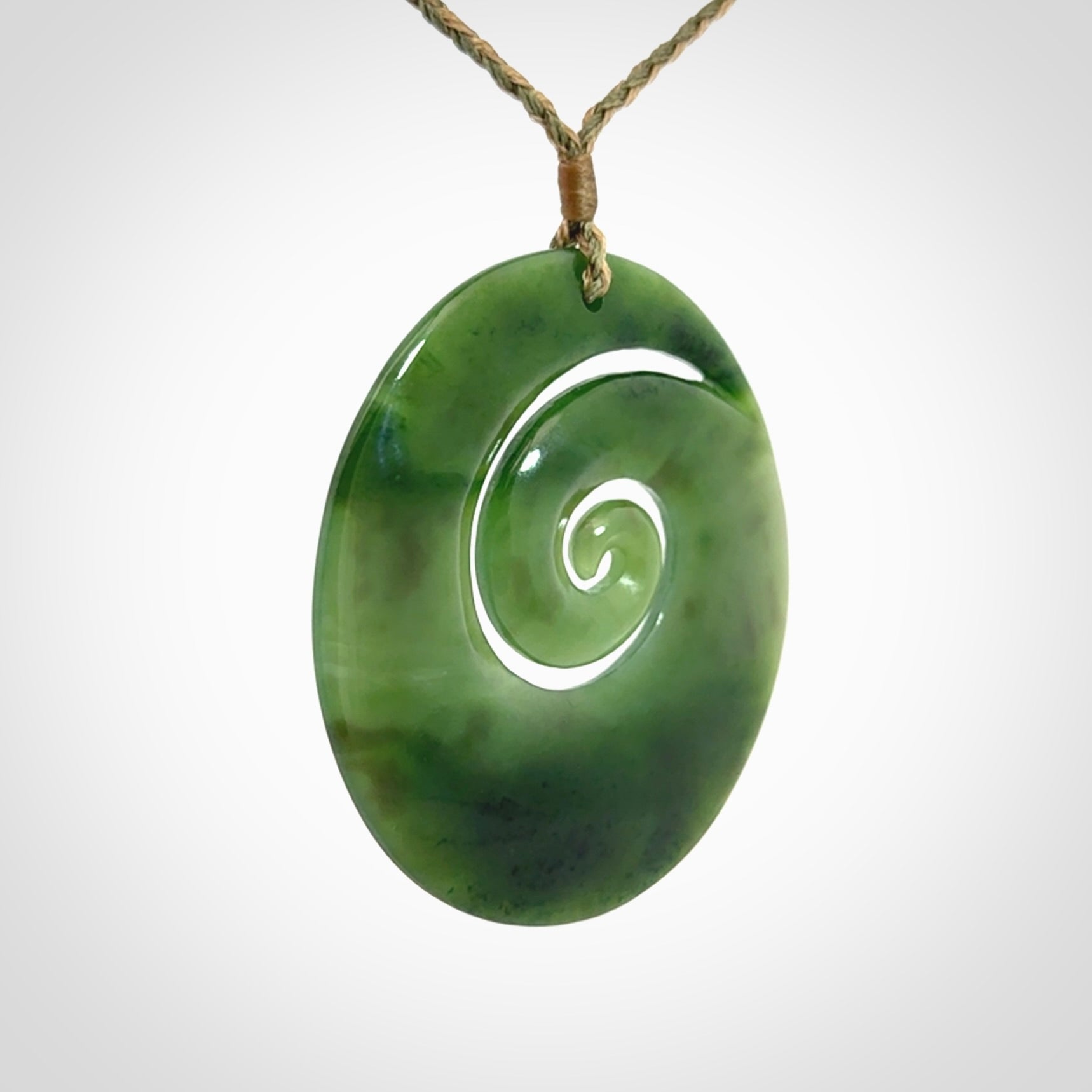 This picture shows a large hand-carved koru. It is carved from British Columbian jade and is suspended from a hand-plaited, tan/green cord.