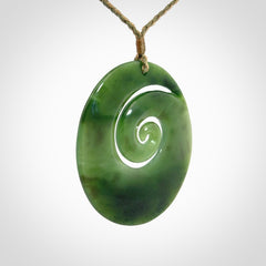 This picture shows a large hand-carved koru. It is carved from British Columbian jade and is suspended from a hand-plaited, tan/green cord.