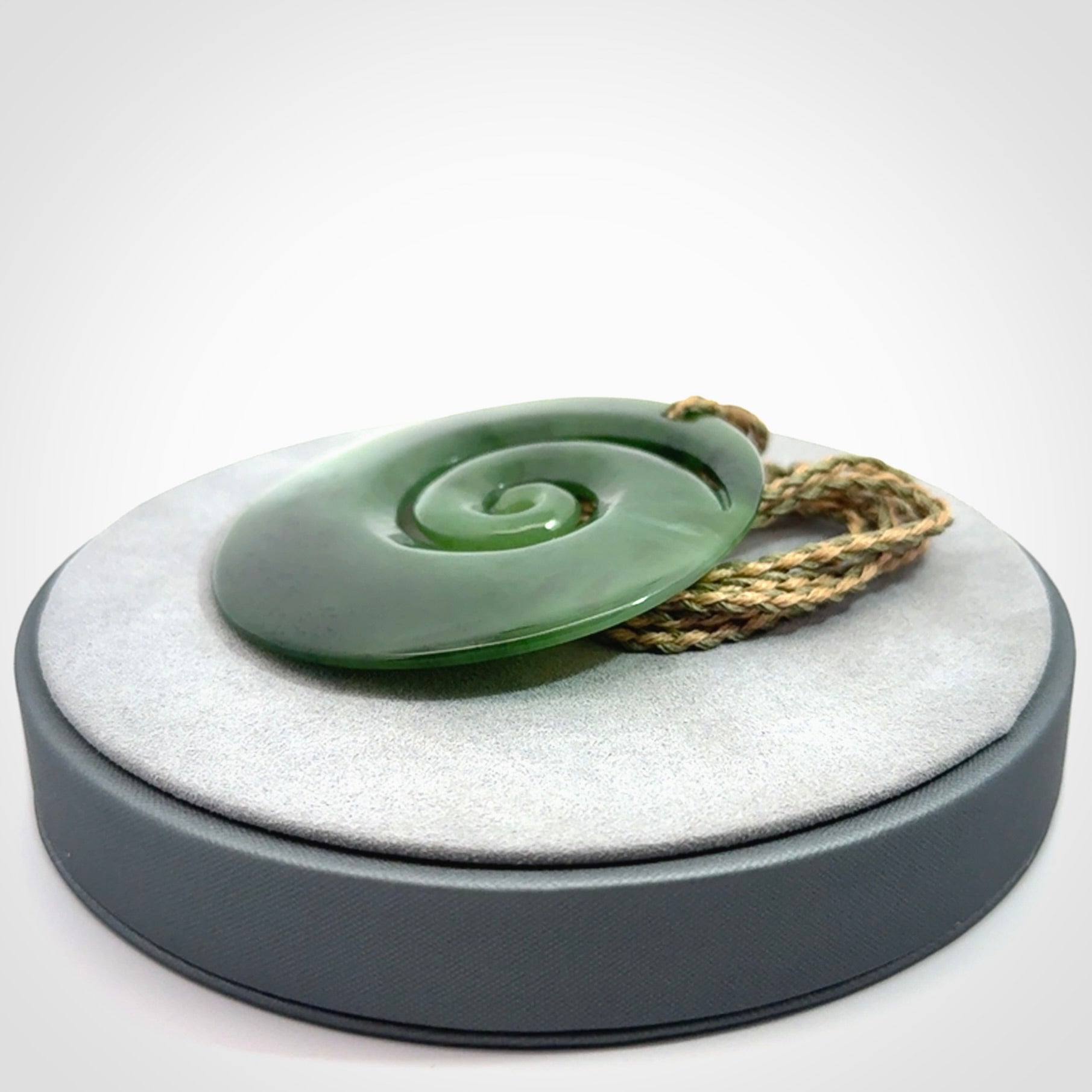 This picture shows a large hand-carved koru. It is carved from British Columbian jade and is suspended from a hand-plaited, tan/green cord.