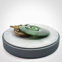 This picture shows a large hand-carved koru. It is carved from British Columbian jade and is suspended from a hand-plaited, tan/green cord.