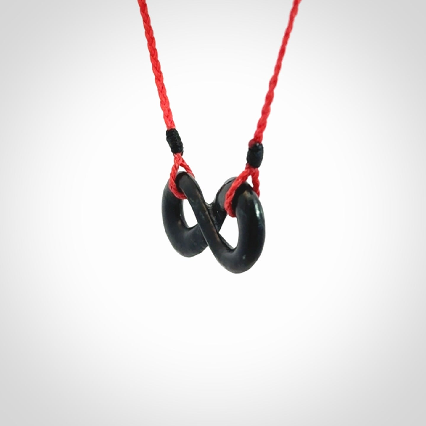 This piece is a fine, delicate eternity shaped pendant. It was carved from a lovely dark piece of black jade. It is suspended on an red plaited cord that is length adjustable.