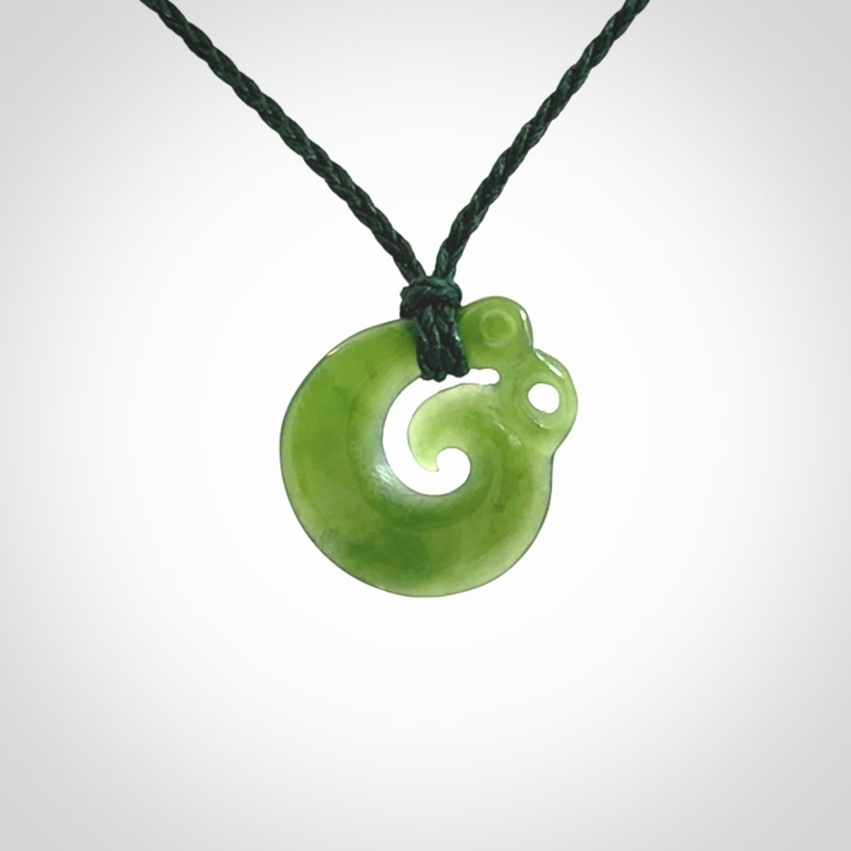 New Zealand Koru Manaia. Hand carved small jade koropepe pendant. Made in New Zealand. Real Jade, pounamu. Provided with an adjustable Manuka Green cord and packaged in a woven kete pouch.