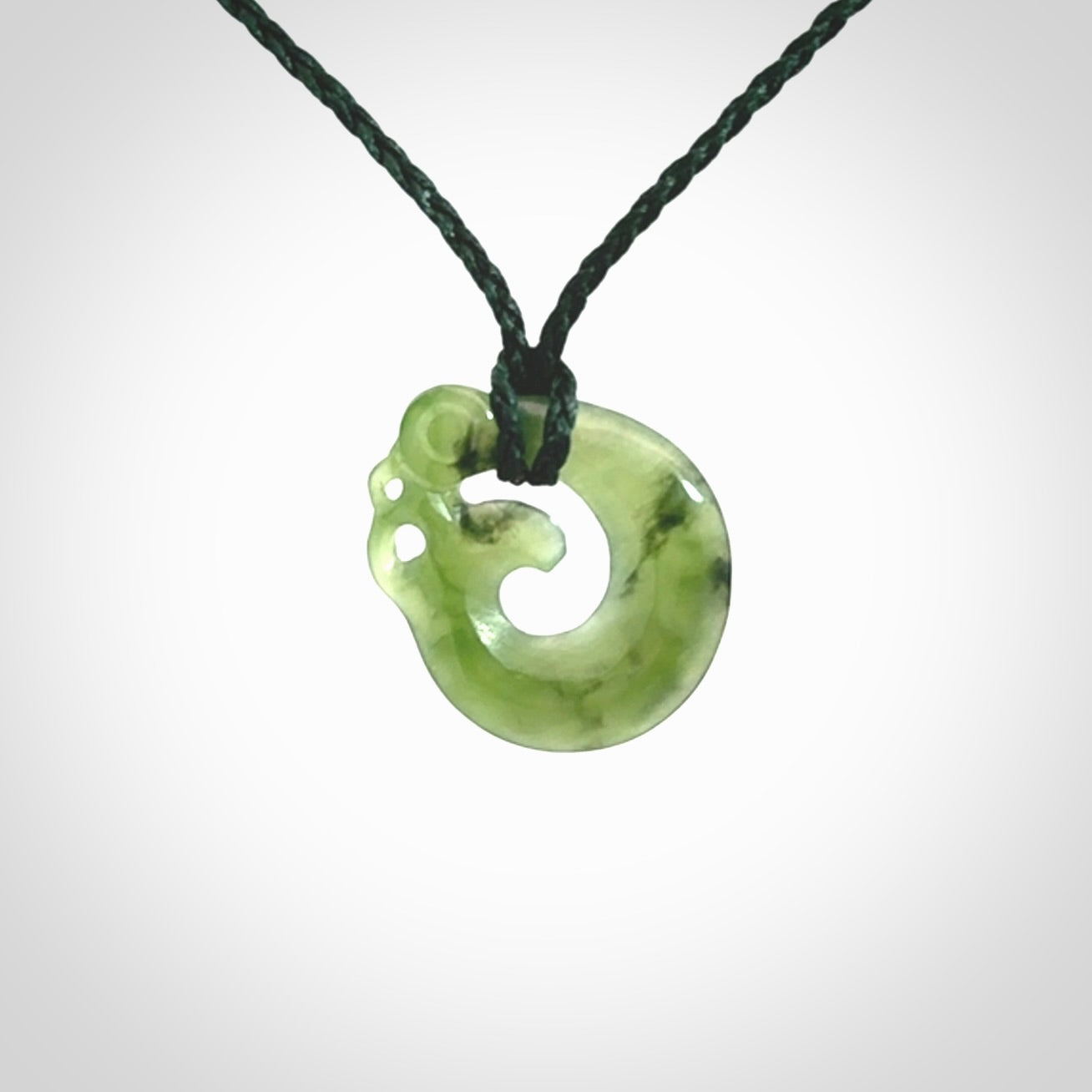 New Zealand Koru Manaia. Hand carved small jade koropepe pendant. Made in New Zealand. Real Jade, pounamu. Provided with an adjustable Manuka Green cord and packaged in a woven kete pouch.