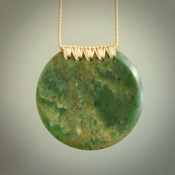 This piece is a large, oval round, disc pendant. It was carved for us by Ric Moor from a lovely deep and milky green piece of New Zealand flower jade. It is suspended on a beige coloured braided cord that is length adjustable.
