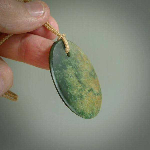 This piece is a large oval round, disc pendant. It was carved for us by Ric Moor from a lovely orange and green piece of New Zealand flower jade. It is suspended on a beige coloured braided cord that is length adjustable.