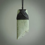 This picture shows a traditional toki pendant carved from New Zealand  jade. The stone is a pale, milky green colour with darker inclusions through it. The cord is a black colour and the binding on the head of the toki is black.