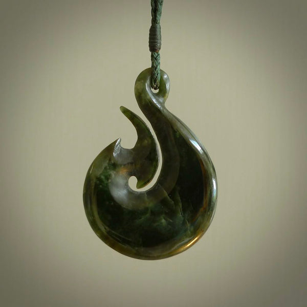 This photo shows a large matau, or hook, pendant carved in deep green New Zealand  Jade. It is a very interesting piece representing a hook. It is strung on a tan cord which is adjustable. The artist who carved this pendant is Ross Crump, a master carver of jade from New Zealand.