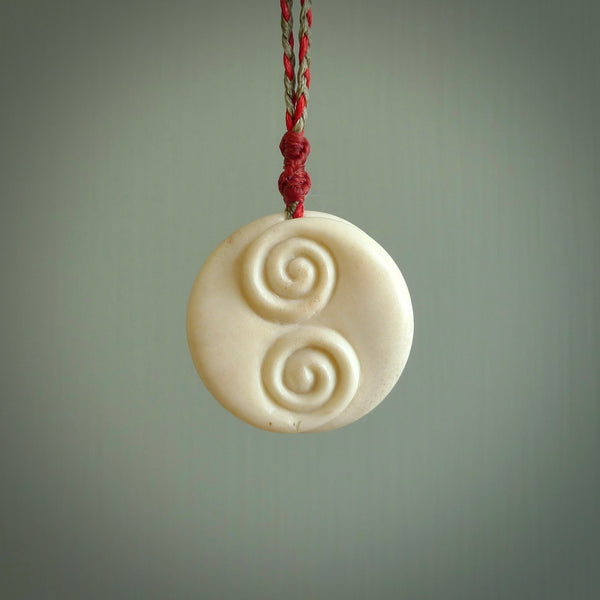 A hand carved and intricate koru pendant made for us by Yuri Terenyi. This is a beautiful little piece and is emblematic of the well known and loved Koru design. It is carved from bone in a round shape with decorative design carved into the koru. It is suspended from a red/khaki cord with a red floret and the necklace is adjustable.