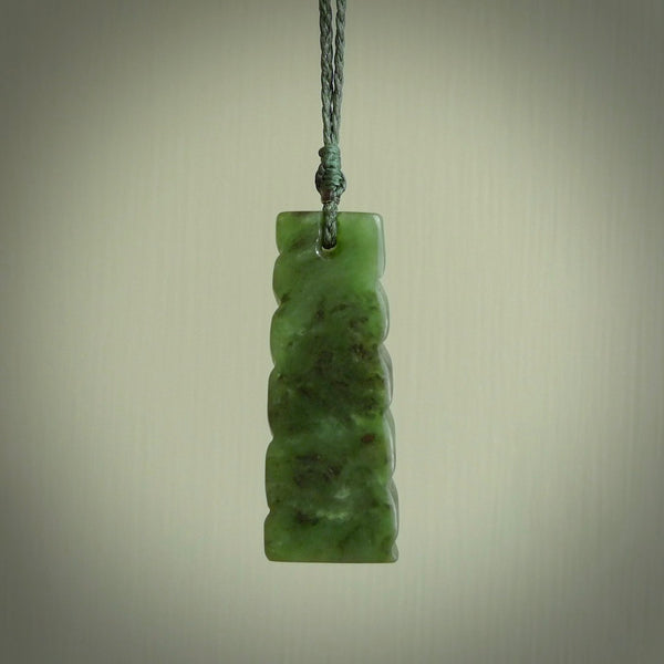 This piece is a toki pendant with a double twist through the body. It is carved from flower jade from New Zealand. It is a deep green colour. The cord is black and is length adjustable.