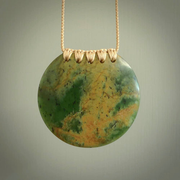 This piece is a large, oval round, disc pendant. It was carved for us by Ric Moor from a lovely deep and milky green piece of New Zealand flower jade. It is suspended on a beige coloured braided cord that is length adjustable.
