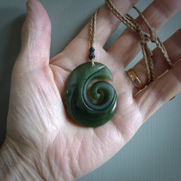 This photo shows a koru with a face carved into the leading edge. The artist, Kerry Thompson, has carved this beautifully and he has polished parts of the carving to a high shine, and other parts he has left in a matte finish. The contrast is beautiful. The pendant is suspended from a plaited Kalahari cord which is adjustable.