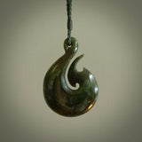 This photo shows a large matau, or hook, pendant carved in deep green New Zealand  Jade. It is a very interesting piece representing a hook. It is strung on a tan cord which is adjustable. The artist who carved this pendant is Ross Crump, a master carver of jade from New Zealand.