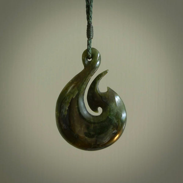 This photo shows a large matau, or hook, pendant carved in deep green New Zealand  Jade. It is a very interesting piece representing a hook. It is strung on a tan cord which is adjustable. The artist who carved this pendant is Ross Crump, a master carver of jade from New Zealand.