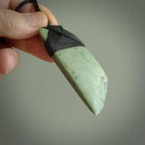 This picture shows a traditional toki pendant carved from New Zealand  jade. The stone is a pale, milky green colour with darker inclusions through it. The cord is a black colour and the binding on the head of the toki is black.