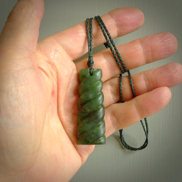 This piece is a toki pendant with a double twist through the body. It is carved from flower jade from New Zealand. It is a deep green colour. The cord is black and is length adjustable.