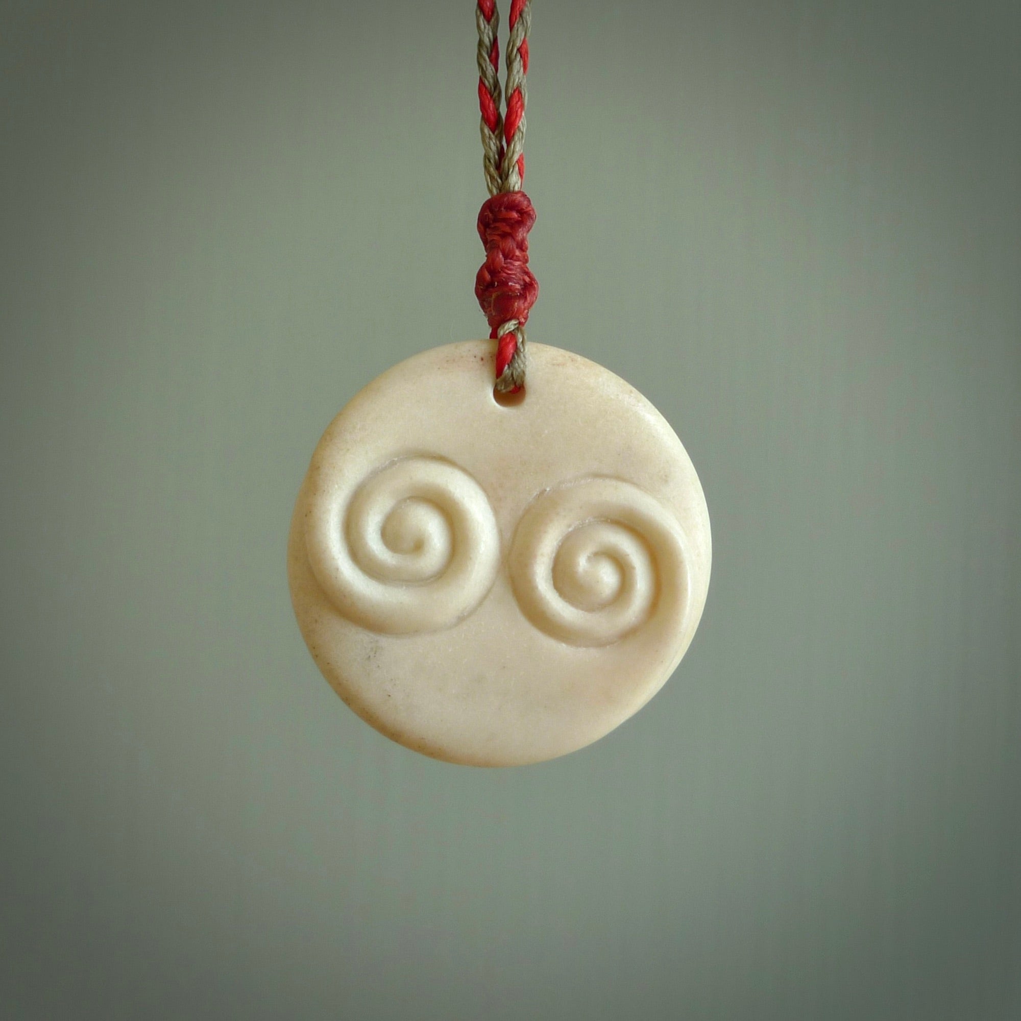 A hand carved and intricate koru pendant made for us by Yuri Terenyi. This is a beautiful little piece and is emblematic of the well known and loved Koru design. It is carved from bone in a round shape with decorative design carved into the koru. It is suspended from a red/khaki cord with a red floret and the necklace is adjustable.