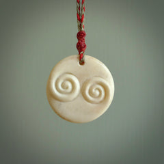 A hand carved and intricate koru pendant made for us by Yuri Terenyi. This is a beautiful little piece and is emblematic of the well known and loved Koru design. It is carved from bone in a round shape with decorative design carved into the koru. It is suspended from a red/khaki cord with a red floret and the necklace is adjustable.