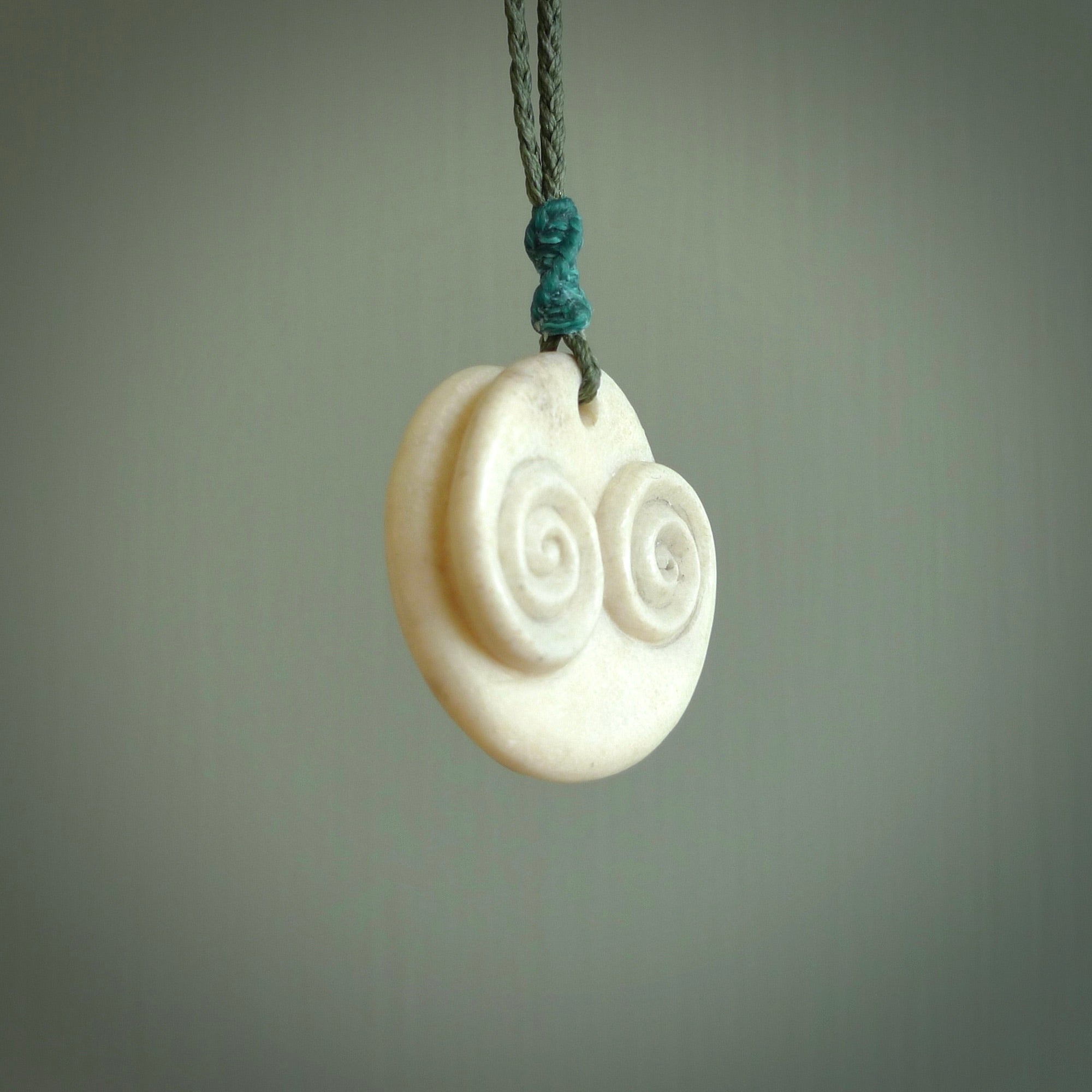A hand carved and intricate koru pendant made for us by Yuri Terenyi. This is a beautiful little piece and is emblematic of the well known and loved Koru design. It is carved from bone in a round shape with decorative design carved into the koru. It is suspended from a sage cord with a paradise blue floret and the necklace is adjustable.
