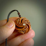 A hand carved and intricate koru pendant made for us by Yuri Terenyi. This is a beautiful little piece and is emblematic of the well known and loved Koru design. It is carved from bone in a hollowed, oval ball shape with decorative design carved into the koru. It is suspended from a Black cord with a wapiti brown floret and the necklace is adjustable.