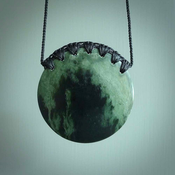 This piece is a large, oval round, disc pendant. It was carved for us by Ric Moor from a lovely deep and milky green piece of New Zealand flower jade. It is suspended on an black coloured braided cord that is length adjustable.