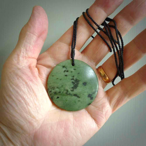 This piece is a large, oval round, disc pendant. It was carved for us by Ric Moor from a lovely deep and milky green piece of New Zealand kokopu jade. It is suspended on a black coloured braided cord that is length adjustable.