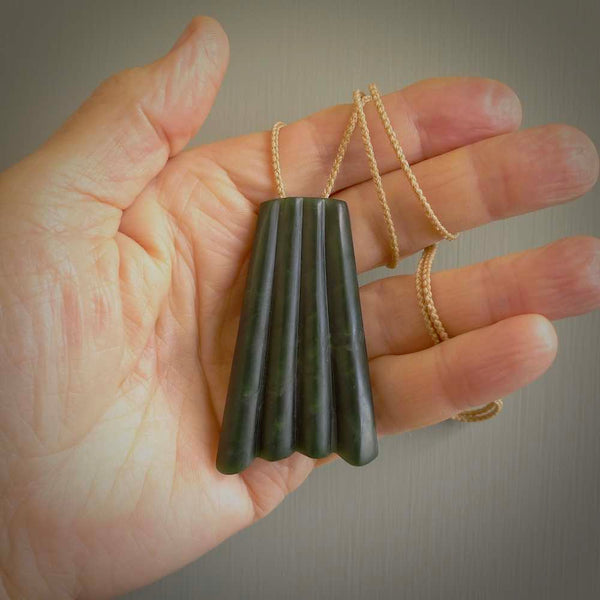 This picture shows a large jade contemporary pendant carved in a deep green coloured piece of marsden jade. This is a unique one off piece. It is suspended on a beige cord which is length adjustable.