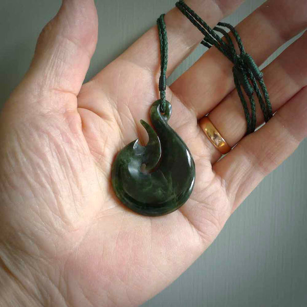 This photo shows a large matau, or hook, pendant carved in deep green New Zealand  Jade. It is a very interesting piece representing a hook. It is strung on a tan cord which is adjustable. The artist who carved this pendant is Ross Crump, a master carver of jade from New Zealand.