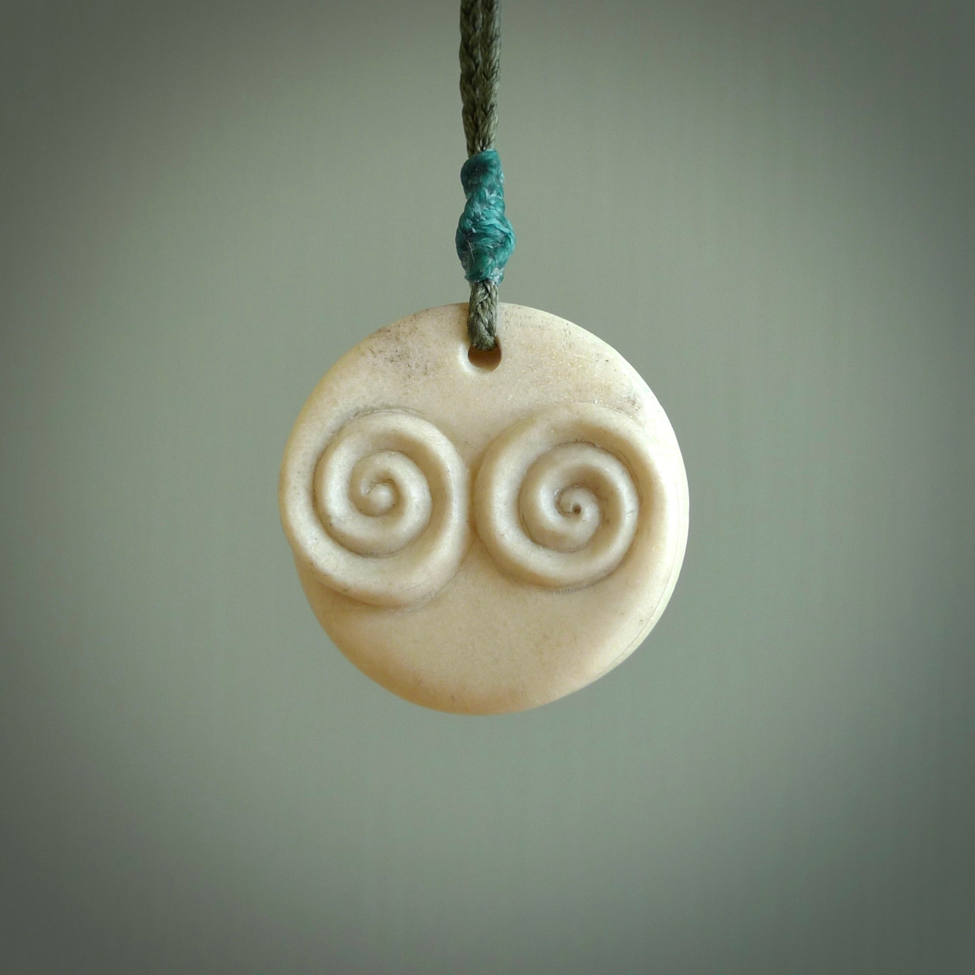 A hand carved and intricate koru pendant made for us by Yuri Terenyi. This is a beautiful little piece and is emblematic of the well known and loved Koru design. It is carved from bone in a round shape with decorative design carved into the koru. It is suspended from a sage cord with a paradise blue floret and the necklace is adjustable.