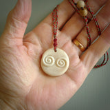 A hand carved and intricate koru pendant made for us by Yuri Terenyi. This is a beautiful little piece and is emblematic of the well known and loved Koru design. It is carved from bone in a round shape with decorative design carved into the koru. It is suspended from a red/khaki cord with a red floret and the necklace is adjustable.