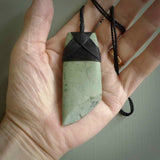 This picture shows a traditional toki pendant carved from New Zealand  jade. The stone is a pale, milky green colour with darker inclusions through it. The cord is a black colour and the binding on the head of the toki is black.