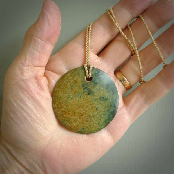 This piece is a large oval round, disc pendant. It was carved for us by Ric Moor from a lovely orange and green piece of New Zealand flower jade. It is suspended on a beige coloured braided cord that is length adjustable.