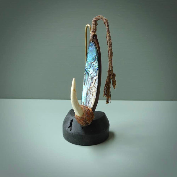 This picture shows a small sculptural hook called a pā kahawai. It is carved from whale bone, paua shell and wood, and is a colourful blue grey colour. It is shown on a stand made from argillite stone and is held in a brass cradle. One only, free shipping worldwide.