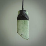 This picture shows a traditional toki pendant carved from New Zealand  jade. The stone is a pale, milky green colour with darker inclusions through it. The cord is a black colour and the binding on the head of the toki is black.
