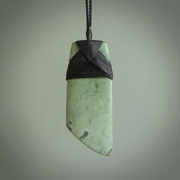 This picture shows a traditional toki pendant carved from New Zealand  jade. The stone is a pale, milky green colour with darker inclusions through it. The cord is a black colour and the binding on the head of the toki is black.