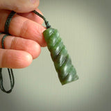 This piece is a toki pendant with a double twist through the body. It is carved from flower jade from New Zealand. It is a deep green colour. The cord is black and is length adjustable.