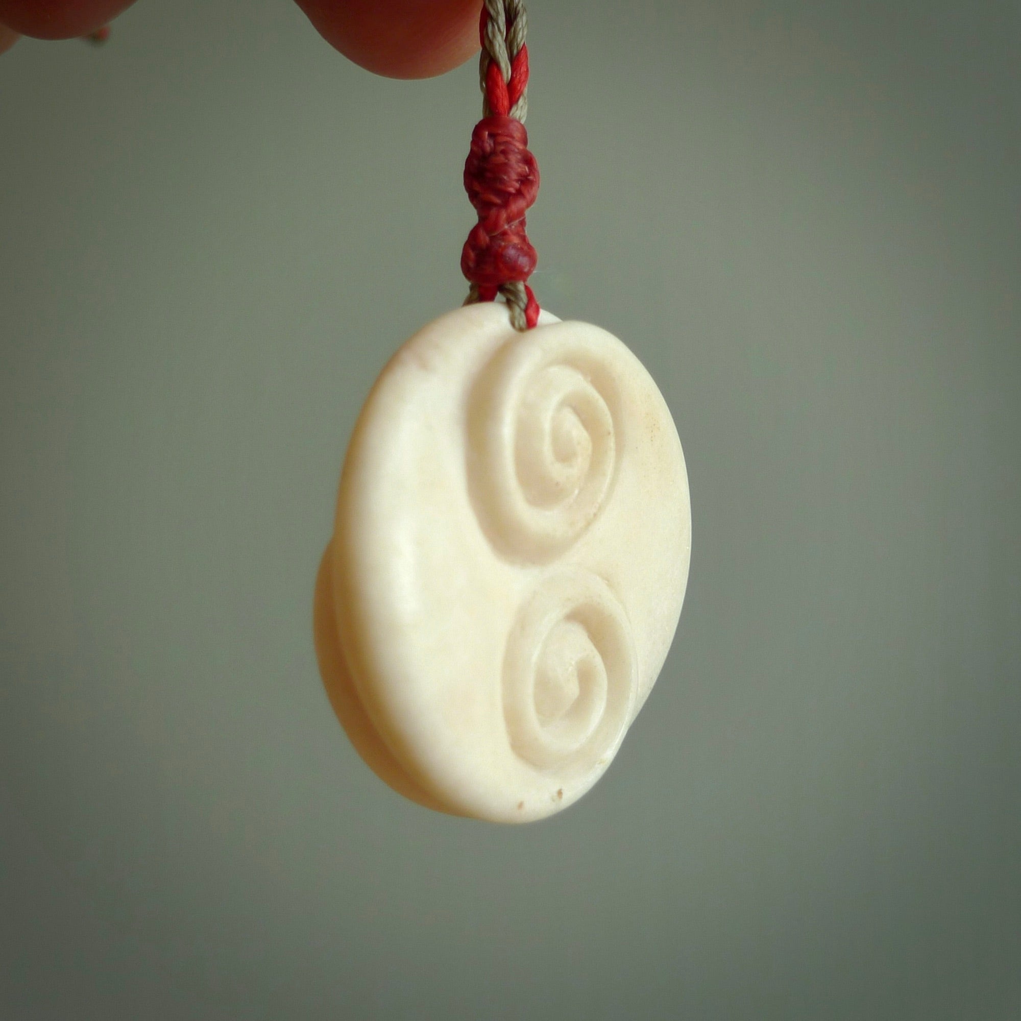 A hand carved and intricate koru pendant made for us by Yuri Terenyi. This is a beautiful little piece and is emblematic of the well known and loved Koru design. It is carved from bone in a round shape with decorative design carved into the koru. It is suspended from a red/khaki cord with a red floret and the necklace is adjustable.