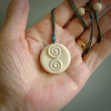 A hand carved and intricate koru pendant made for us by Yuri Terenyi. This is a beautiful little piece and is emblematic of the well known and loved Koru design. It is carved from bone in a round shape with decorative design carved into the koru. It is suspended from a sage cord with a paradise blue floret and the necklace is adjustable.
