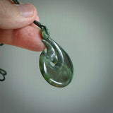This photo shows a large matau, or hook, pendant carved in deep green New Zealand  Jade. It is a very interesting piece representing a hook. It is strung on a tan cord which is adjustable. The artist who carved this pendant is Ross Crump, a master carver of jade from New Zealand.