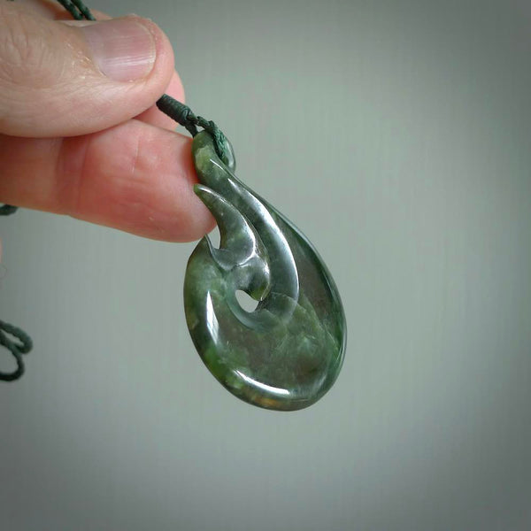 This photo shows a large matau, or hook, pendant carved in deep green New Zealand  Jade. It is a very interesting piece representing a hook. It is strung on a tan cord which is adjustable. The artist who carved this pendant is Ross Crump, a master carver of jade from New Zealand.