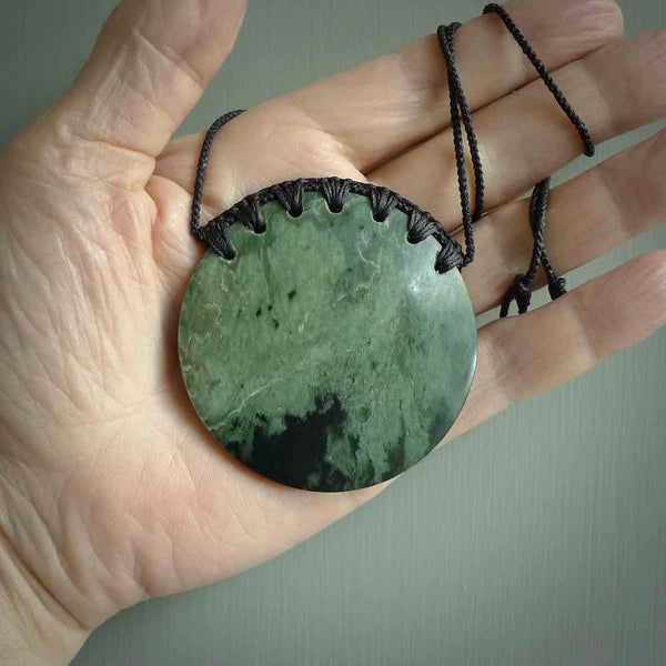 This piece is a large, oval round, disc pendant. It was carved for us by Ric Moor from a lovely deep and milky green piece of New Zealand flower jade. It is suspended on an black coloured braided cord that is length adjustable.