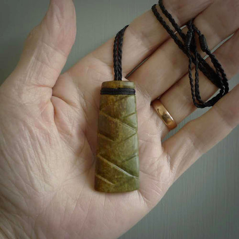 This picture shows a toki pendant carved with a woven front face and a cupped back. It is made from a piece of rare New Zealand Flower Jade by our artist and carver Ana Krakosky. The cord is black and is length adjustable.