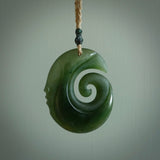 This photo shows a koru with a face carved into the leading edge. The artist, Kerry Thompson, has carved this beautifully and he has polished parts of the carving to a high shine, and other parts he has left in a matte finish. The contrast is beautiful. The pendant is suspended from a plaited Kalahari cord which is adjustable.