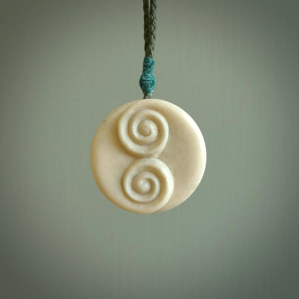 A hand carved and intricate koru pendant made for us by Yuri Terenyi. This is a beautiful little piece and is emblematic of the well known and loved Koru design. It is carved from bone in a round shape with decorative design carved into the koru. It is suspended from a sage cord with a paradise blue floret and the necklace is adjustable.