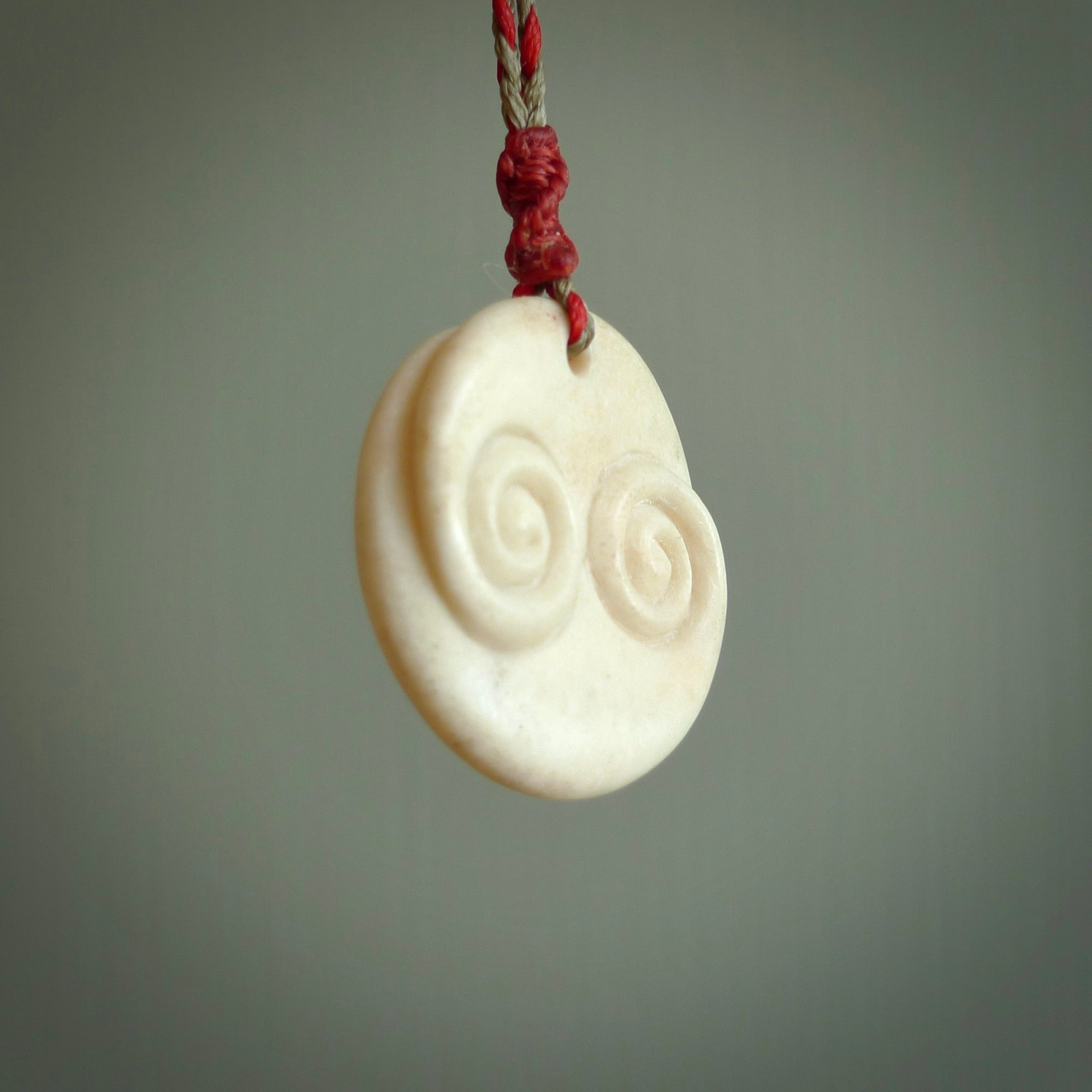 A hand carved and intricate koru pendant made for us by Yuri Terenyi. This is a beautiful little piece and is emblematic of the well known and loved Koru design. It is carved from bone in a round shape with decorative design carved into the koru. It is suspended from a red/khaki cord with a red floret and the necklace is adjustable.