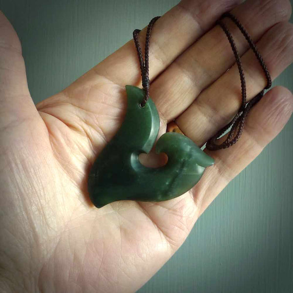 This photo shows a perfectly New Zealand jade hook pendant. It is hand carved from a deep green piece of New Zealand jade and is suspended on a brown four plait cord that is length adjustable.