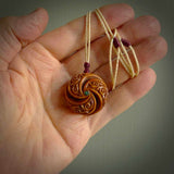 A hand carved and intricate koru pendant made for us by Yuri Terenyi. This is a beautiful little piece and is emblematic of the well known and loved Koru design. It is carved from bone in a hollowed, oval ball shape with decorative design carved into the koru. It is suspended from an Ice White cord with a burgundy floret and the necklace is adjustable.