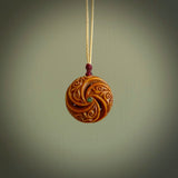 A hand carved and intricate koru pendant made for us by Yuri Terenyi. This is a beautiful little piece and is emblematic of the well known and loved Koru design. It is carved from bone in a hollowed, oval ball shape with decorative design carved into the koru. It is suspended from an Ice White cord with a burgundy floret and the necklace is adjustable.