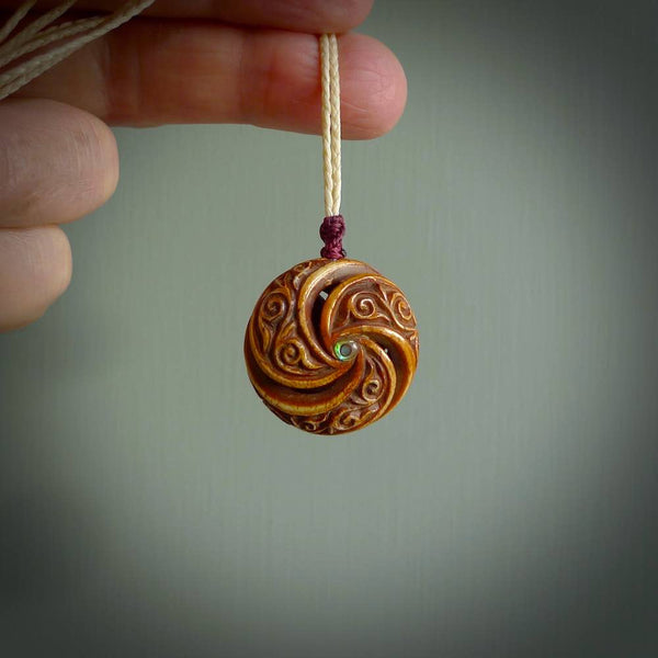 A hand carved and intricate koru pendant made for us by Yuri Terenyi. This is a beautiful little piece and is emblematic of the well known and loved Koru design. It is carved from bone in a hollowed, oval ball shape with decorative design carved into the koru. It is suspended from an Ice White cord with a burgundy floret and the necklace is adjustable.